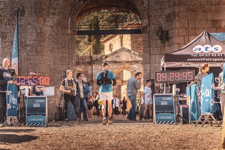 Arrivée d'un trail sous un porche en pierre, horloges digitales et tente de chronométrage ATS-Sport
